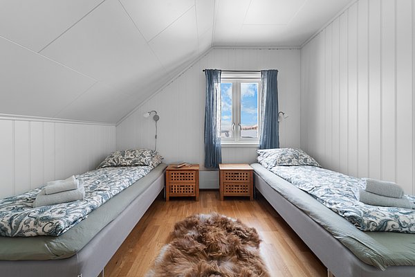 Gemütliches Schlafzimmer in Nord-Norwegen mit zwei Einzelbetten, Dachschräge, Holzboden und Fenster mit blauem Vorhang. Ein Fellteppich zwischen den Betten sorgt für rustikales Flair. Ideal für entspannte Angelurlaube.