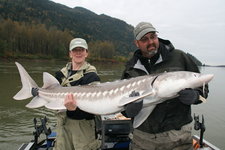 Zwei Personen halten gemeinsam einen großen Stör vor einer malerischen Flusslandschaft.