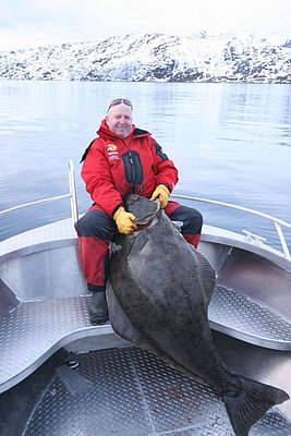 Ein Angler hält einen großen Heilbutt auf einem Boot vor schneebedeckten Bergen in Nord-Norwegen. Perfektes Ziel für Angelreisen mit beeindruckenden Fangmöglichkeiten. Ideal für Abenteuer und entspannte Angeltage.