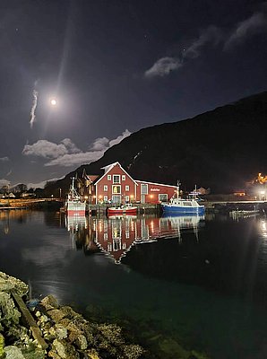 Im Mondlicht spiegelt sich ein rotes Hafengebäude in einem ruhigen Fjord. Fischerboote liegen am Pier, umgeben von Bergen – perfekte Kulisse für Angelreisen in Südwest-Norwegen.