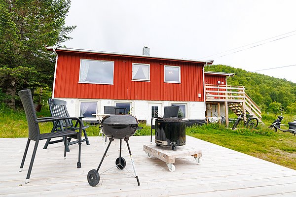 Rotes Holzhaus in Nord-Norwegen mit Terrasse und Gartenmöbeln, umgeben von grüner Landschaft; idealer Ausgangspunkt für Angelurlaub und Naturerlebnisse. Perfekt für erholsame Tage in idyllischer Umgebung.