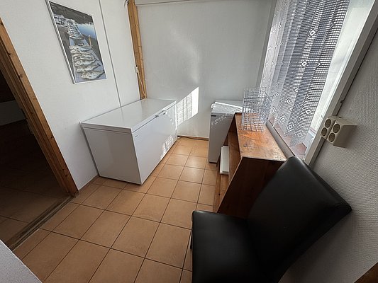 Kleiner, heller Raum in Hitra, Norwegen, mit Gefriertruhe, Stuhl, Wandbild mit Fischmotiv und Sonnenschein durch Spitzenvorhänge, ideal für Angler.