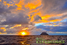 Sonnenuntergang über dem Meer in Tansania, mit dramatischen Wolken am Himmel und einer silhouettierten Insel. Perfekt für einen Angelurlaub, der Abenteuerlust und Entspannung vereint. Ideal für Reisende, die unberührte Natur lieben.