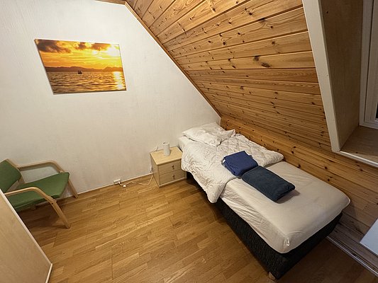 Gemütliches Schlafzimmer mit Holzwänden auf Hitra, Norwegen. Einzelbett mit frischer Bettwäsche, dekorativem Sonnenuntergangsbild und kleinem Nachttisch. Ideal für entspannte Angelreisen in malerischer Umgebung.