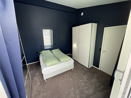 Gemütliches Schlafzimmer mit dunkelblauen Wänden auf Hitra, Norwegen. Doppelbett mit grüner Bettwäsche, weißem Schrank und kleinem Fenster. Ideal für erholsame Nächte nach einem erfolgreichen Angelurlaub.