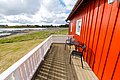 Rotes, idyllisches Ferienhaus auf Hitra, Norwegen. Holzterrasse mit zwei Stühlen und Tisch, Blick auf Wiese und Küste. Perfekt für Angelurlaub und Erholung in atemberaubender Natur. Ideal für erholsame Angelreisen in malerischer Umgebung.