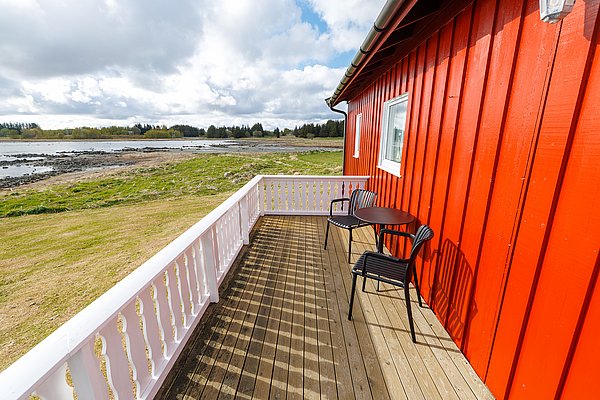 Rotes, idyllisches Ferienhaus auf Hitra, Norwegen. Holzterrasse mit zwei Stühlen und Tisch, Blick auf Wiese und Küste. Perfekt für Angelurlaub und Erholung in atemberaubender Natur. Ideal für erholsame Angelreisen in malerischer Umgebung.
