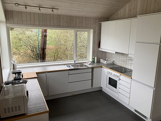 Moderne, helle Küche in Hitra, Norwegen, mit großen Fenstern, die einen Blick auf grüne Natur bieten; schlichte weiße Schränke, Elektrogeräte und viel Arbeitsfläche; idealer Rückzugsort nach einem Angelabenteuer.