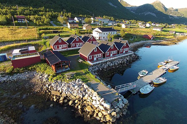 Luftaufnahme von roten Fischerhütten am Fjord in Nord-Norwegen. Der Steg mit Booten lädt zum Angeln ein, umgeben von grünen Bergen und malerischer Natur. Ideal für erholsame Angelurlaube.