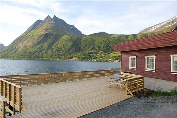Atemberaubendes Panorama in Nordland, Norwegen: Traditionelles rotes Holzhaus mit Terrasse am Fjord vor majestätischen Bergen. Ideal für Angelreisen und Naturliebhaber. Entdecke die idyllische Ruhe und grandiose Landschaft für einen unvergesslichen Angelurlaub.