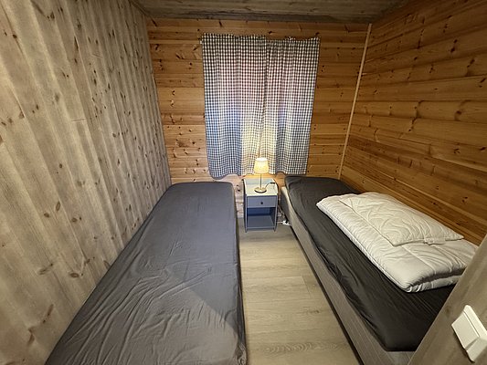 Gemütliches Schlafzimmer in einer Holzhütte auf Hitra, Norwegen. Zwei Einzelbetten, gestreifte Vorhänge und warme Beleuchtung schaffen eine einladende Atmosphäre. Ideal für Angelurlauber und Naturliebhaber, die Entspannung und Abenteuer suchen.