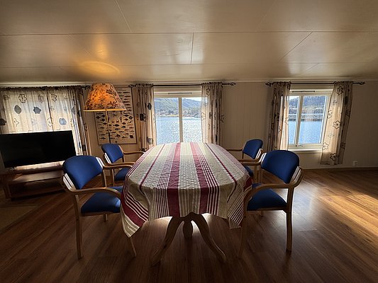 Gemütliches Wohnzimmer in Hitra, Norwegen, mit Tisch, blauen Stühlen und großen Fenstern, die einen herrlichen Blick auf das Meer bieten. Ideal für Angelurlauber, die Komfort und atemberaubende Landschaften suchen.