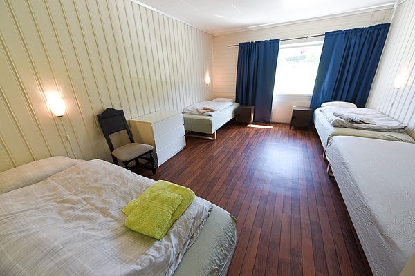 Gemütliches Schlafzimmer in Hitra, Norwegen, mit vier Einzelbetten, Holzboden, sanfter Beleuchtung und blauen Vorhängen. Ideal für Angelurlauber, die Erholung und Komfort nach einem erfolgreichen Angeltag suchen.