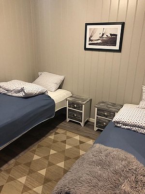 Gemütliches Schlafzimmer in Nord-Norwegen mit zwei Einzelbetten, blauen Decken und Nachttischen. An der Wand hängt ein Schwarz-Weiß-Bild eines Segelboots. Ideal für Angelurlauber, die Komfort und Erholung suchen.