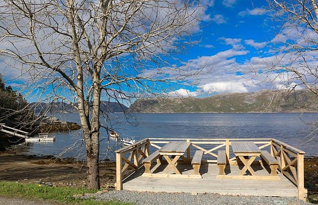 Holzterrasse mit Picknicktischen an der Küste von Hitra, Norwegen. Blick auf ruhiges Wasser und bewaldete Hügel im Hintergrund, ideal für Angelurlaub und Naturerlebnis. Klarer Himmel und friedliche Stimmung.