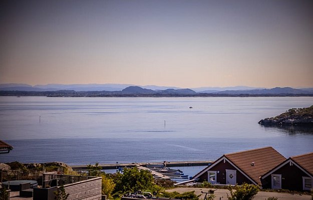 Küstenlandschaft in Südwest-Norwegen: Ruhiges Meer, Holzhäuser am Ufer, malerische Fjorde und Hügel im Hintergrund. Perfekt für einen Angelurlaub mit atemberaubender Naturkulisse zur Entspannung und Abenteuer.