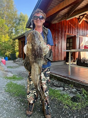 Person hält großen Fisch vor rustikalem Holzhaus in Hitra, Norwegen. Perfekter Angelurlaub für Abenteurer und Naturfreunde, umgeben von malerischer Landschaft. Optimal für unvergessliche Angelerlebnisse und Entspannung in der Natur.