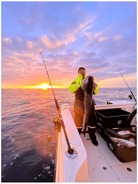 Eine Person in auffälliger Kleidung hält einen großen Fisch auf einem Boot bei Sonnenuntergang, während Angelruten auf das ruhige Meer zeigen.