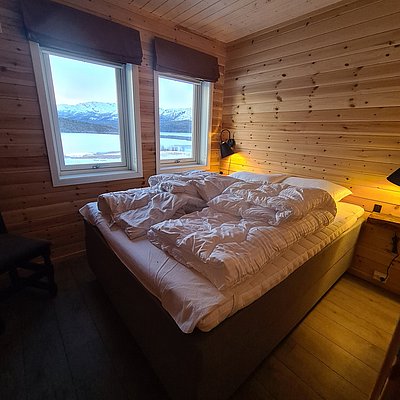 Gemütliches Schlafzimmer in einer skandinavischen Hütte mit Holzwänden. Großes Fenster bietet Blick auf die schneebedeckte Landschaft Nord-Norwegens. Ideal für erholsamen Angelurlaub und Abenteuer in der Natur.