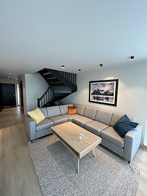 Ein stilvoll eingerichteter Wohnbereich in Nordland, Norwegen, mit grauem Sofa, Holztisch, moderner Treppe und Landschaftsfotografie an der Wand. Perfekt für entspannte Angelreisen mit Komfort.