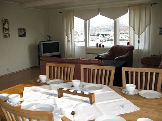Gemütliches Wohnzimmer in Nordland, Norwegen, mit Holztisch, gedeckt für sechs Personen, Ledersofas und Blick auf den Hafen durch ein großes Fenster. Ideal für entspannte Angelurlaube in idyllischer Lage.