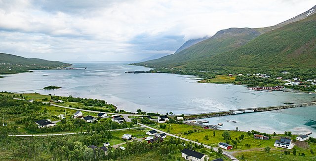 Luftaufnahme einer idyllischen Küstenlandschaft in Nord-Norwegen. Ein ruhiger Fjord umgeben von sattgrünen Bergen und verstreuten Häusern. Ideal für Angelabenteuer und Naturliebhaber. Über eine Brücke gelangt man in die unberührte Natur.