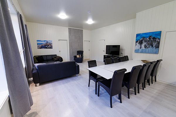 Gemütlicher Aufenthaltsraum in Nordland, Norwegen: Moderne Einrichtung mit schwarzem Sofa, großem Esstisch, TV und stimmungsvollen Gemälden an den Wänden. Ideal für Entspannung nach erlebnisreichen Angelabenteuern.
