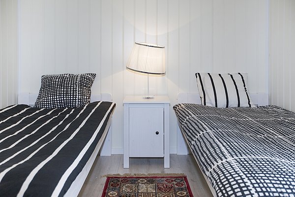 Gemütliches Schlafzimmer in Nord-Norwegen. Zwei Einzelbetten mit schwarz-weiß karierten und gestreiften Bettbezügen, weiße Wände und Nachttischlampe. Ideal für entspannende Angelurlaube. Skandinavisches Design trifft Komfort.