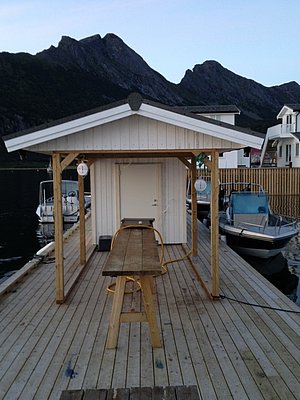 Kleines Holzgebäude auf einem Pier in Nordland, Norwegen. Umgeben von Booten, ruhig gelegen vor einer imposanten Bergkulisse. Perfekt für Angelabenteuer und Erkundung der atemberaubenden Natur beim Angelurlaub.
