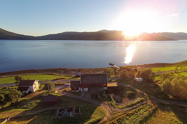 Sonnenaufgang über einem ruhigen, glitzernden Fjord in Nord-Norwegen. Traditionelle Holzhäuser und grüne Wiesen im Vordergrund strahlen Ruhe und Naturschönheit aus. Perfekt für Angelurlaub und Entspannung in unberührter Landschaft.