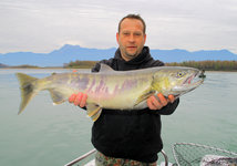 Eine Person hält einen großen Fisch, vermutlich ein Lachs, mit einer Berglandschaft und einem ruhigen See im Hintergrund.