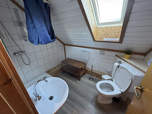 Helles Badezimmer in Hitra, Norwegen mit Dachfenster, weißer Holzverkleidung und blauen Duschvorhängen. Ausstattung: Dusche, WC, Waschbecken und Holzregal. Ideal für Angelurlaub mit Komfort.