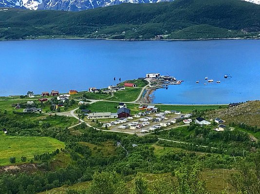 Küstenlandschaft in Nord-Norwegen: Malerisches Dorf am Fjord mit bunten Häusern, Wohnmobilen und Booten im Hafen. Umgeben von grünen Hügeln und schneebedeckten Bergen – ein Paradies für Angelreisen und Naturerlebnisse.