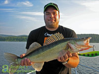 Ein Angler hält einen gefangenen Zander vor einem malerischen See im Hintergrund in die Kamera.