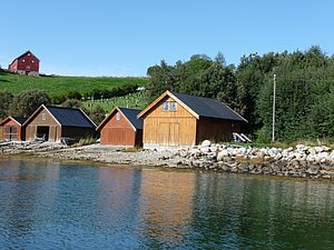 Rote und braune Holzhütten am klaren Wasser in Hitra, Norwegen. Grüne Wiesen und Bäume im Hintergrund. Die ruhige Umgebung lädt ein zum Angelurlaub und Entspannen in der Natur.