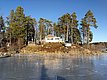Kleines weißes Haus auf einem Hügel, umgeben von hohen Bäumen, mit gefrorenem See im Vordergrund und blauem Himmel im Hintergrund.