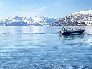 Ein Boot gleitet über einen klaren, spiegelnden Fjord in Nord-Norwegen, umgeben von schneebedeckten Bergen. Perfekt für Angelreisen und Naturerlebnisse in arktischer Umgebung.