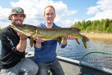 Zwei lächelnde Personen in einem Boot halten gemeinsam einen gefangenen Hecht vor einer malerischen Seenlandschaft.
