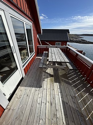 Rote Holzhütte auf Hitra, Norwegen, mit Holzterrasse und Meerblick. Blauer Himmel und felsige Küste im Hintergrund. Ideal für Angelreisen und entspannte Urlaubstage in skandinavischer Natur.