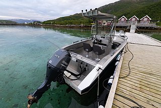 Ein modernes Angelboot liegt an einem Holzsteg in Nord-Norwegen. Kristallklares Wasser, umgeben von grünen Bergen und roten Hütten, lädt zum Abenteuer ein. Ideal für Angelreisen und Naturerlebnisse.