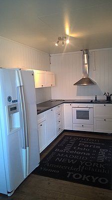 Moderne, weiße Küche in Nord-Norwegen mit großem Kühlschrank, schwarzer Arbeitsfläche und stilvollem Ofen; der Bodenläufer trägt Städtenamen. Perfekt für Selbstversorger im Angelurlaub.
