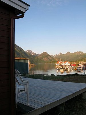 Sonnenuntergang in Nordland, Norwegen: Blick von einer Holzterrasse auf ein ruhiges Hafenbecken mit Fischerbooten und beeindruckender Bergkulisse. Ideal für Angelreisen und Naturerlebnisse.