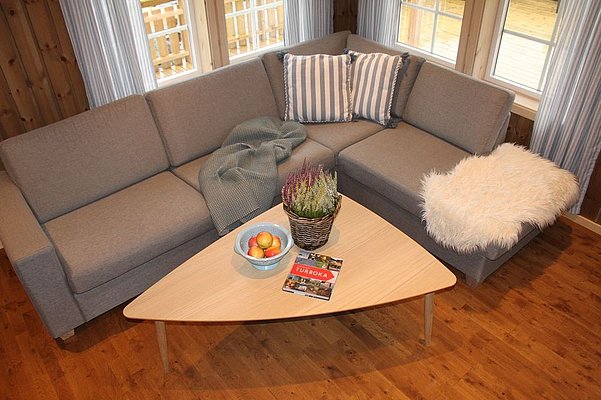 Gemütliches Wohnzimmer in Nordnorwegen mit grauem Ecksofa, Holztisch, Früchteschale und Dekoaccessoires. Ideal für Entspannung nach einem erfolgreichen Angelurlaub. Helle Fenster bieten Blick auf norwegische Landschaft.