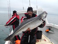 Eine fröhliche Person steht auf einem Boot und hält einen großen Fisch, während eine andere Person im Hintergrund angelt.