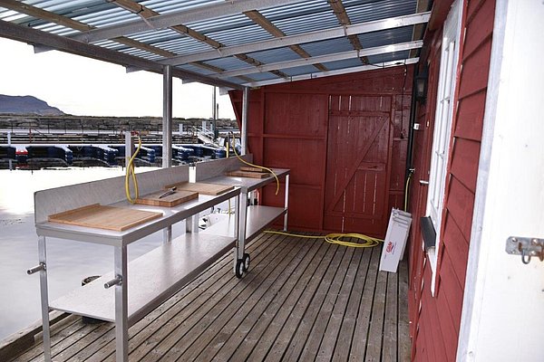 Überdachter Filetierplatz am Wasser in Südwest-Norwegen, ideal für Angler. Rote Holzwände, robuste Arbeitsflächen mit Schneidebrettern und Schläuchen. Perfekt für den Angelurlaub und Frischfisch-Vorbereitung. Abenteuerliche Küstenumgebung.