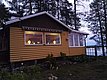 Gemütliches Holzhaus am Waldrand mit beleuchteten Fenstern bei Dämmerung, umgeben von hohen Bäumen.