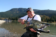 Ein Angler hält lächelnd einen großen Lachs am Flussufer mit bewaldeten Hügeln im Hintergrund.