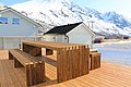 Moderne Holzterrasse in Nord-Norwegen mit Sitzgelegenheiten und beeindruckendem Bergpanorama im Hintergrund. Perfekter Ort für entspannte Momente im Angelurlaub, eingebettet in die Schönheit der schneebedeckten Landschaft.
