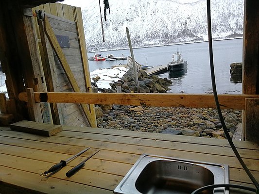 Rustikale Bootsverankerung in Nord-Norwegen: Eine schneebedeckte Küstenlandschaft mit Anlegestelle und Booten. Holzveranda mit Arbeitsfläche und Spüle bietet Aussicht auf verschneite Berge am ruhigen Fjord. Ideal für Angelurlaub und Naturerlebnisse.