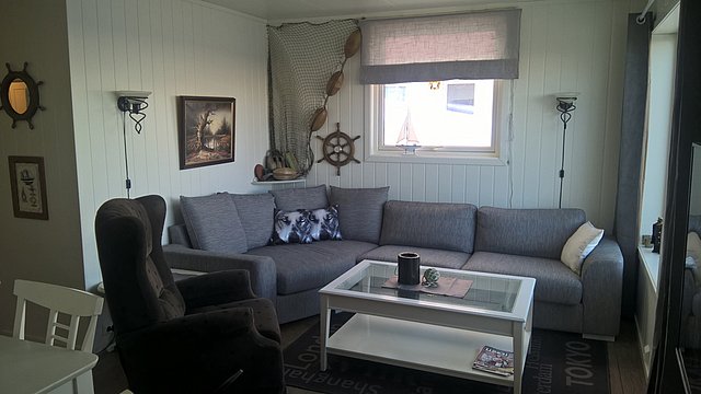 Gemütliches Wohnzimmer in Nord-Norwegen mit grauem Sofa, maritimer Dekoration und hellem Interieur. Perfekte Unterkunft für Angelreisen und entspannende Auszeiten nach erlebnisreichen Tagen auf See. Familienfreundlich und ideal für Naturfreunde.